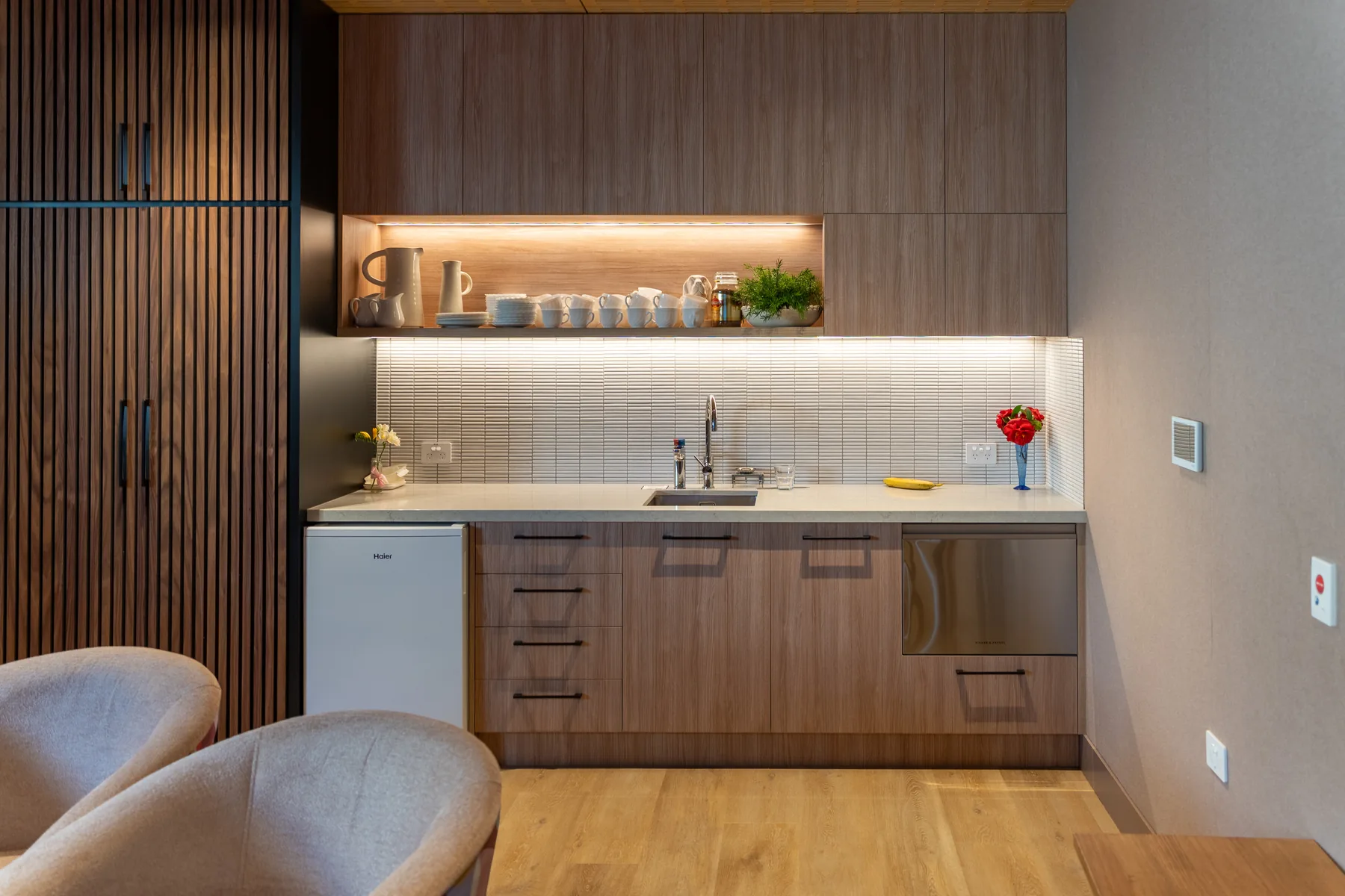 A kitchenette with mugs on an open shelf, a tiled splashback and two armchairs alongside