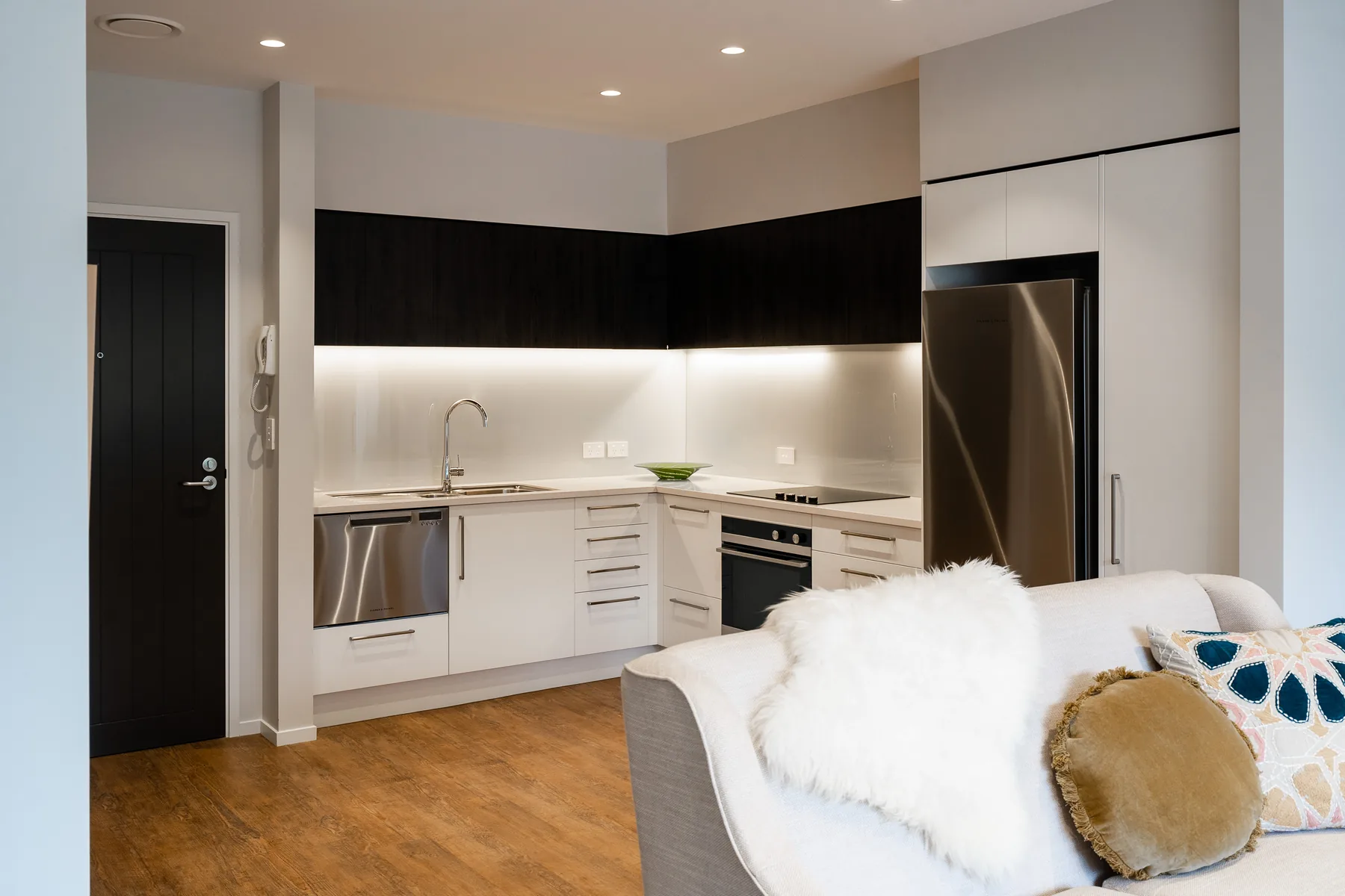 An apartment kitchen with dark upper cabinets, under-cabinet lighting, a built-in oven and a tall fridge-freezer