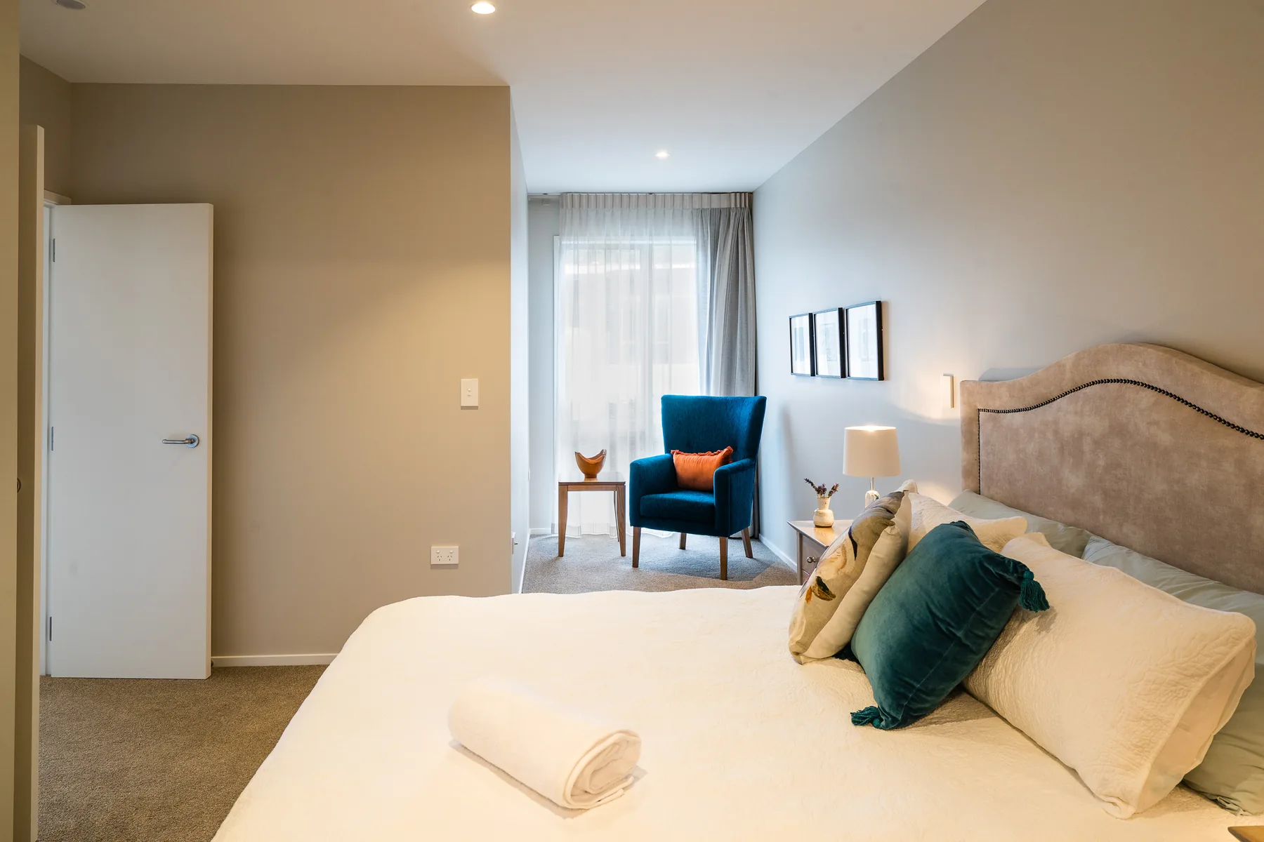 An apartment bedroom with a double bed and upholstered headboard, and a blue armchair beside a curtained window