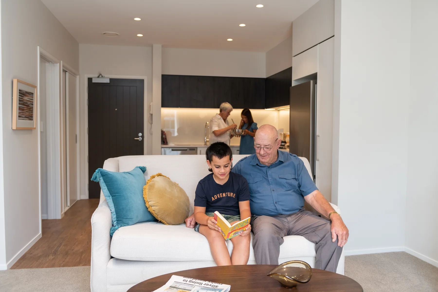 A resident and his young grandson sitting on the sofa reading a book together in an apartment living room, with family in the kitchen behind